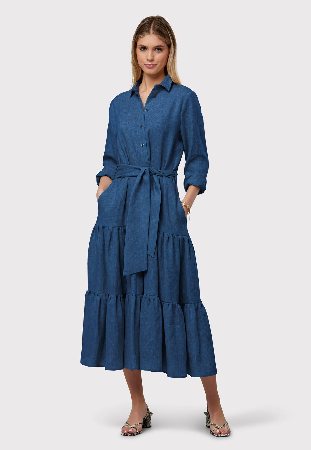Adele Borage Blue Dress, crafted in a rich borage blue linen. The design showcases a classic shirt collar and a buttoned finish at the waist, ensuring a flattering silhouette that accentuates the hips. Transitioning seamlessly into a gathered tiered skirt, it exudes feminine charm. Complete with a detachable belt, practical pockets, and standard full-length shirt sleeves, this dress is versatile and perfect for any occasion.