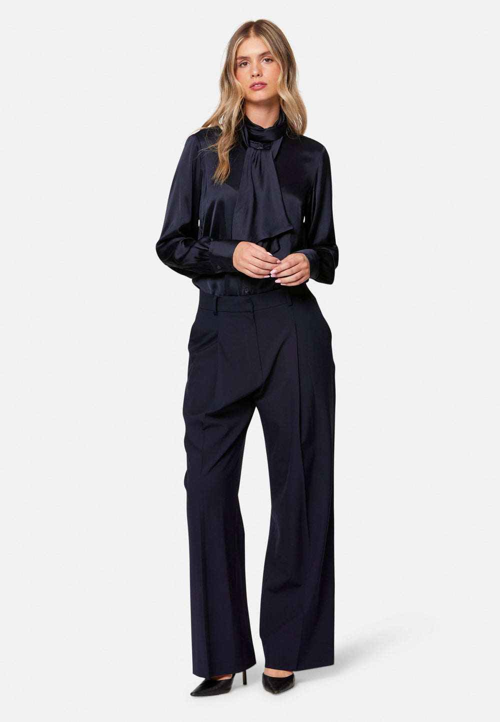 Woman wearing a navy outfit with a blouse with a bow-tie and wide-leg pants on a white background