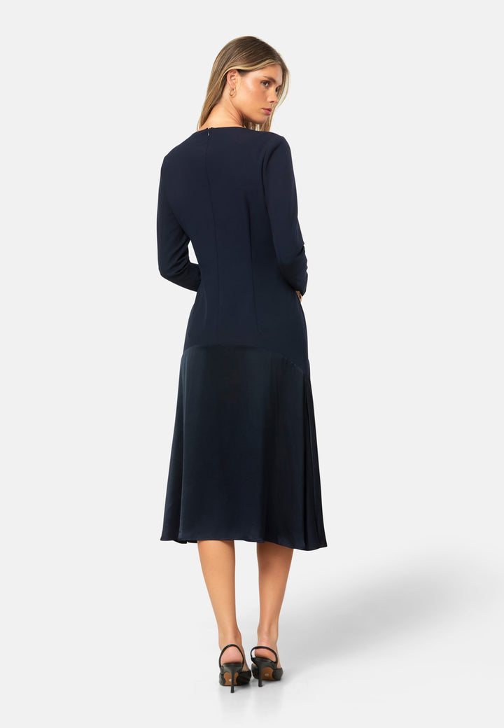Kristy, a modern take on the dropped waist. Cut from plain-weave worsted navy fabric with a touch of stretch, the dress contrasts with a fluid viscose satin skirt that flows softly from the low hip. Designed with a clean round neckline, long fitted sleeves, a dropped waistline and a gently flared midi skirt that falls to mid-calf. The tonal fabric pairing creates subtle dimension, while the detachable satin scarf/belt can be cinched at the waist or draped elegantly around the neck. 