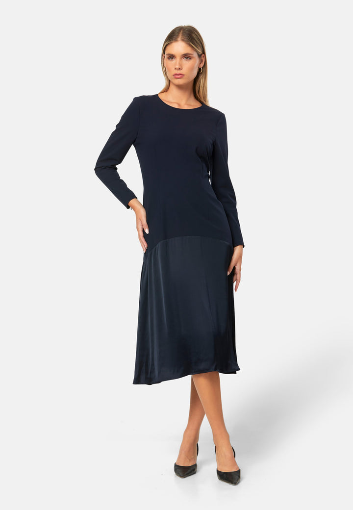 Kristy, a modern take on the dropped waist. Cut from plain-weave worsted navy fabric with a touch of stretch, the dress contrasts with a fluid viscose satin skirt that flows softly from the low hip. Designed with a clean round neckline, long fitted sleeves, a dropped waistline and a gently flared midi skirt that falls to mid-calf. The tonal fabric pairing creates subtle dimension, while the detachable satin scarf/belt can be cinched at the waist or draped elegantly around the neck. 