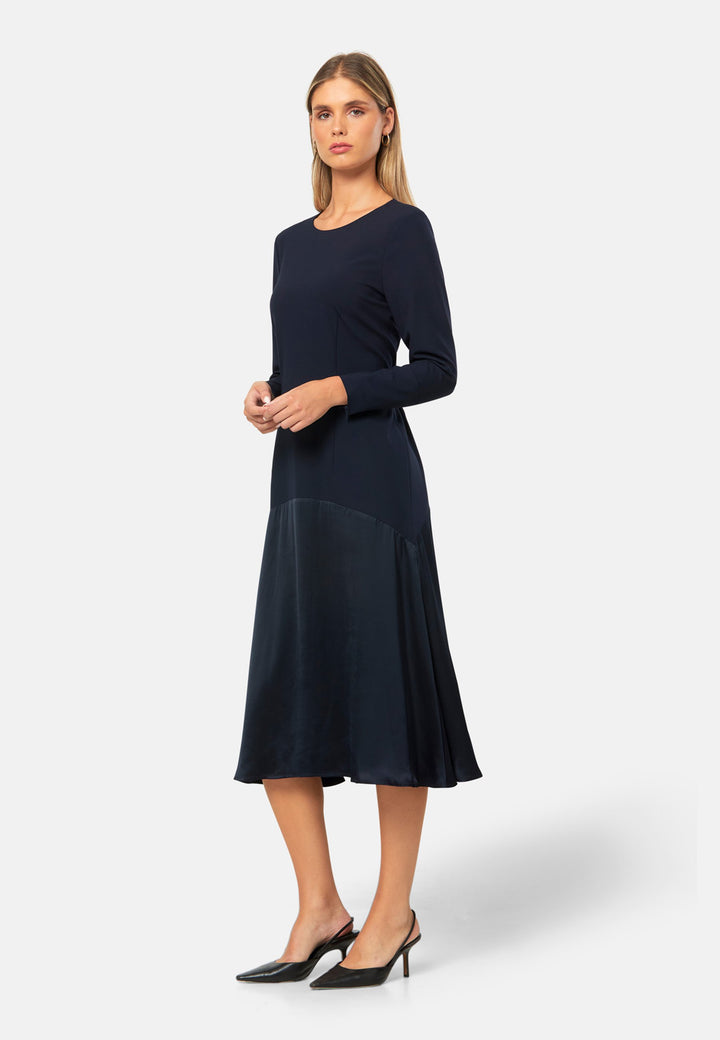 Kristy, a modern take on the dropped waist. Cut from plain-weave worsted navy fabric with a touch of stretch, the dress contrasts with a fluid viscose satin skirt that flows softly from the low hip. Designed with a clean round neckline, long fitted sleeves, a dropped waistline and a gently flared midi skirt that falls to mid-calf. The tonal fabric pairing creates subtle dimension, while the detachable satin scarf/belt can be cinched at the waist or draped elegantly around the neck. 