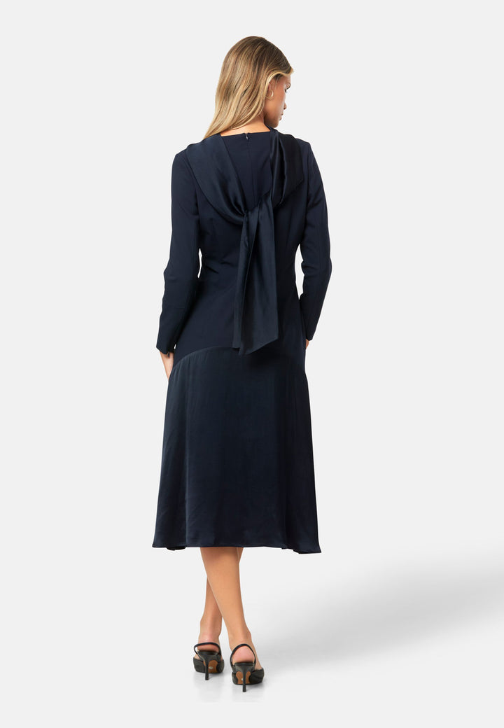 Kristy, a modern take on the dropped waist. Cut from plain-weave worsted navy fabric with a touch of stretch, the dress contrasts with a fluid viscose satin skirt that flows softly from the low hip. Designed with a clean round neckline, long fitted sleeves, a dropped waistline and a gently flared midi skirt that falls to mid-calf. The tonal fabric pairing creates subtle dimension, while the detachable satin scarf/belt can be cinched at the waist or draped elegantly around the neck. 