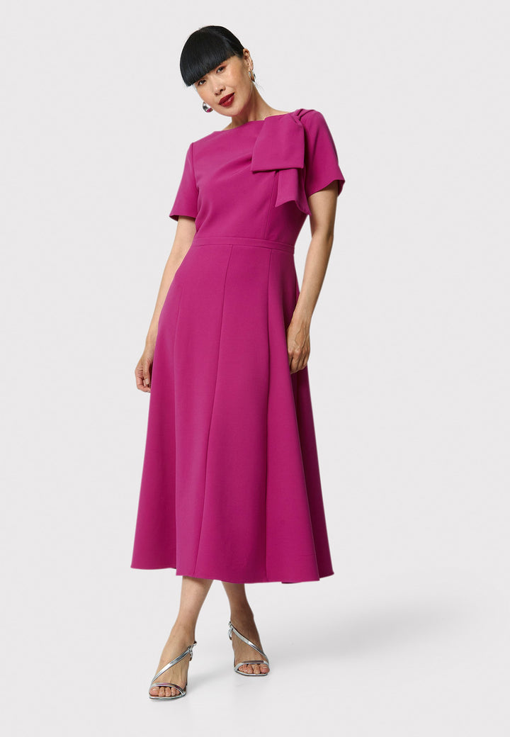 Introducing the Laoise pink dress, a refined statement piece for special occasions. Featuring short sleeves, a fitted bodice, and an elegant shoulder bow detail, this dress has a flattering A-line shape with a cascading paneled skirt. Designed with side seam pockets and a back zip for ease, it combines comfort and sophistication, making it perfect for races, weddings and formal events.