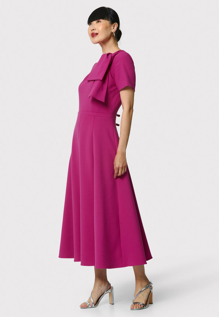 Introducing the Laoise pink dress, a refined statement piece for special occasions. Featuring short sleeves, a fitted bodice, and an elegant shoulder bow detail, this dress has a flattering A-line shape with a cascading paneled skirt. Designed with side seam pockets and a back zip for ease, it combines comfort and sophistication, making it perfect for races, weddings and formal events.
