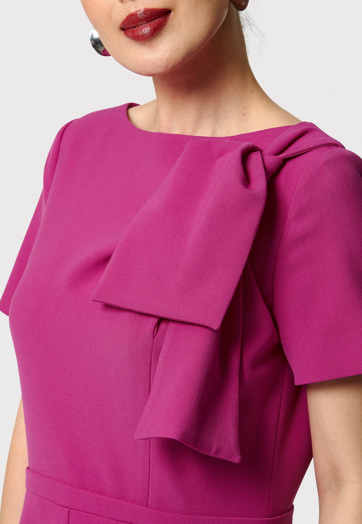 Introducing the Laoise pink dress, a refined statement piece for special occasions. Featuring short sleeves, a fitted bodice, and an elegant shoulder bow detail, this dress has a flattering A-line shape with a cascading paneled skirt. Designed with side seam pockets and a back zip for ease, it combines comfort and sophistication, making it perfect for races, weddings and formal events.