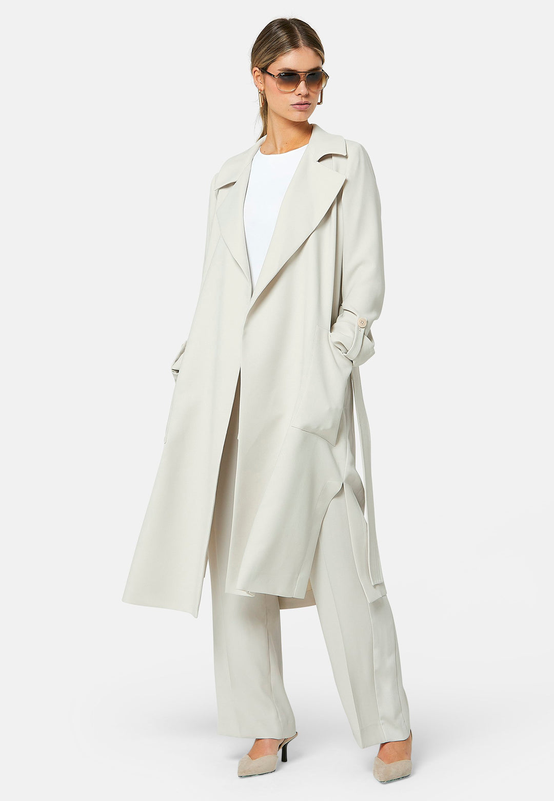 Lydiah, stone trench coat in a fluid satin back crepe. This coat features a relaxed, flowing silhouette, a self-tie belt, and front pockets. The design includes buttoned cuffs and a wide lapel. Pair with coordinating trouser for a chic monochromatic look.