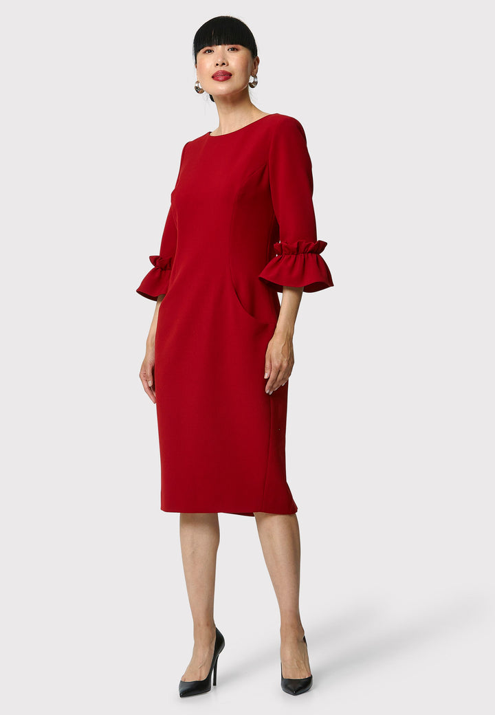 The Maisie Garnet Red Dress, impeccably tailored from signature tricotine fabric with a hint of stretch. This body-skimming silhouette features a round neck and bracelet-length sleeves complemented by exaggerated frill cuffs, ideal for evening cocktails, a night at the theatre, or a stylish winter wedding.