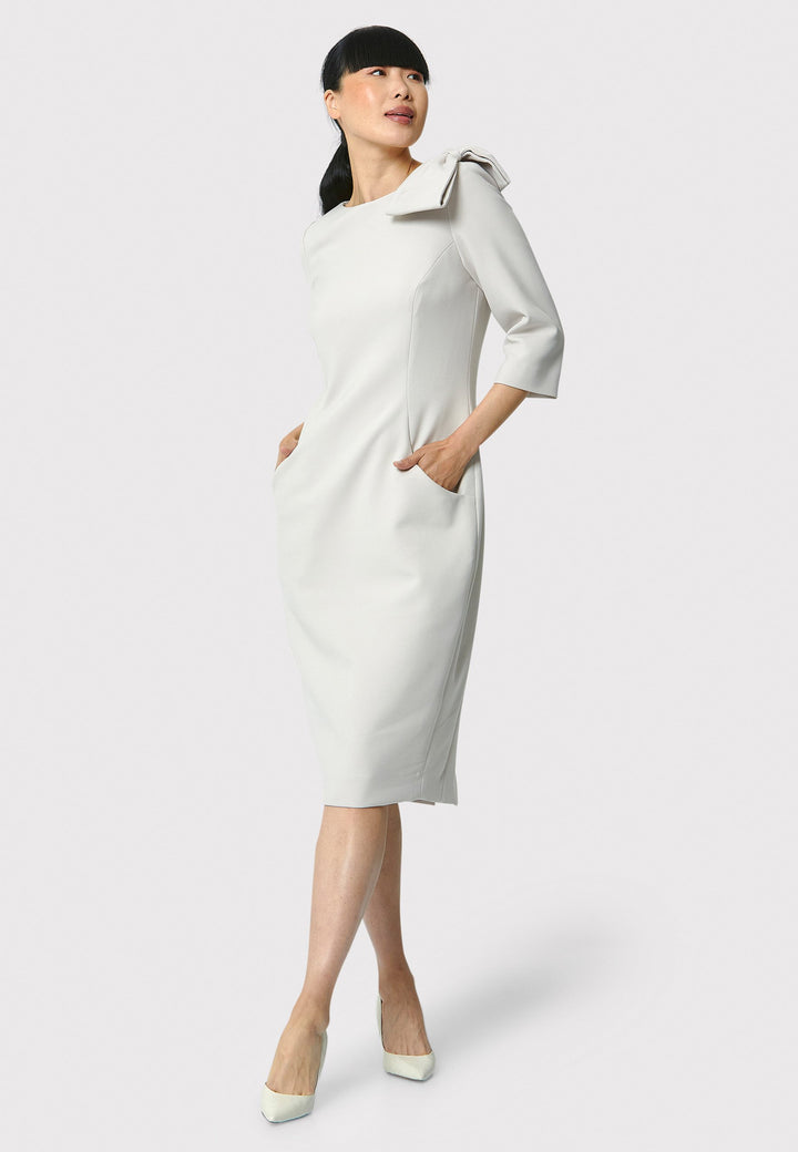 The Natalie dress, crafted from a stone hued tricotine fabric with a hint of stretch, this figure-hugging dress showcases a chic slash neckline, bow detailing, and a mid-calf pencil skirt. Suitable for a night out with friends, a romantic date, or a winter wedding.