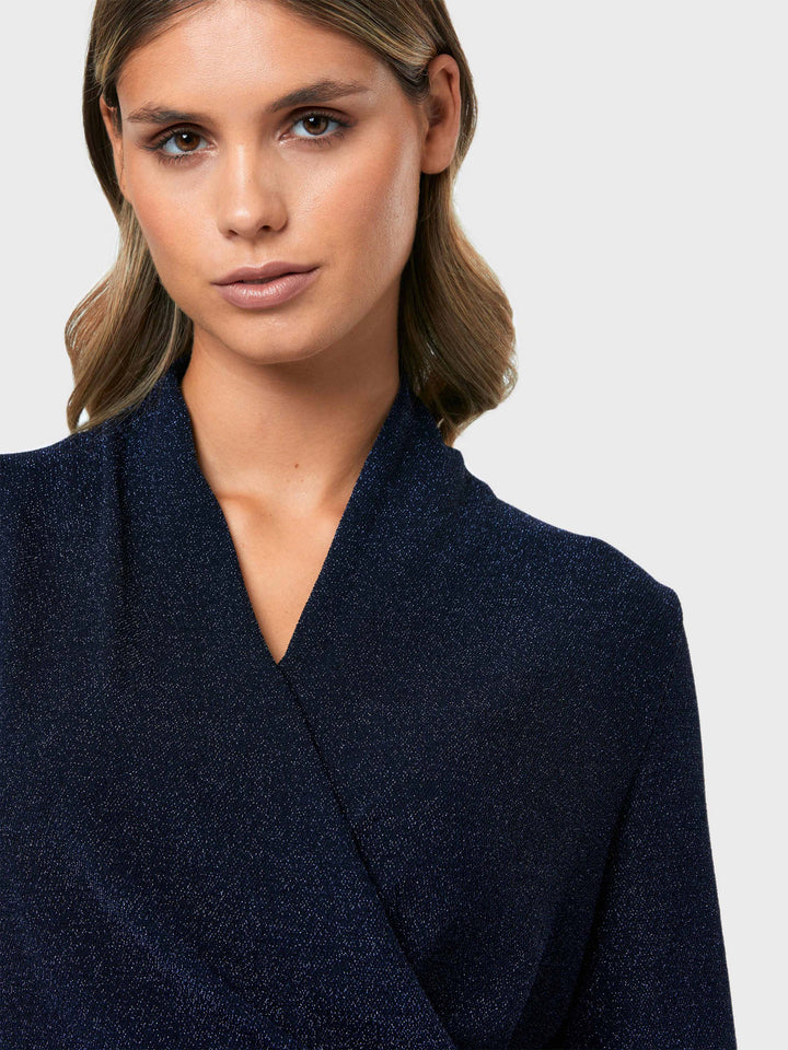 Introducing the Roxanne Midnight Shimmer Top, an updated version of the beloved best-selling faux-wrap top. This season's design features a beautifully draped neckline for an elegant touch. With its hip-length cut and full-length sleeves, this top combines comfort and chic style effortlessly. Crafted from a navy sparkly jersey fabric, it adds a touch of glamour to any outfit. Embrace the ultimate in comfortable chic with the Roxanne Midnight Shimmer Top.