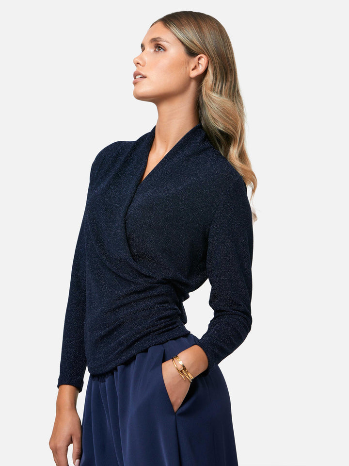 Introducing the Roxanne Midnight Shimmer Top, an updated version of the beloved best-selling faux-wrap top. This season's design features a beautifully draped neckline for an elegant touch. With its hip-length cut and full-length sleeves, this top combines comfort and chic style effortlessly. Crafted from a navy sparkly jersey fabric, it adds a touch of glamour to any outfit. Embrace the ultimate in comfortable chic with the Roxanne Midnight Shimmer Top.