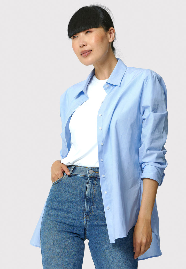 Person wearing a light blue shirt over a white top and jeans on a plain background