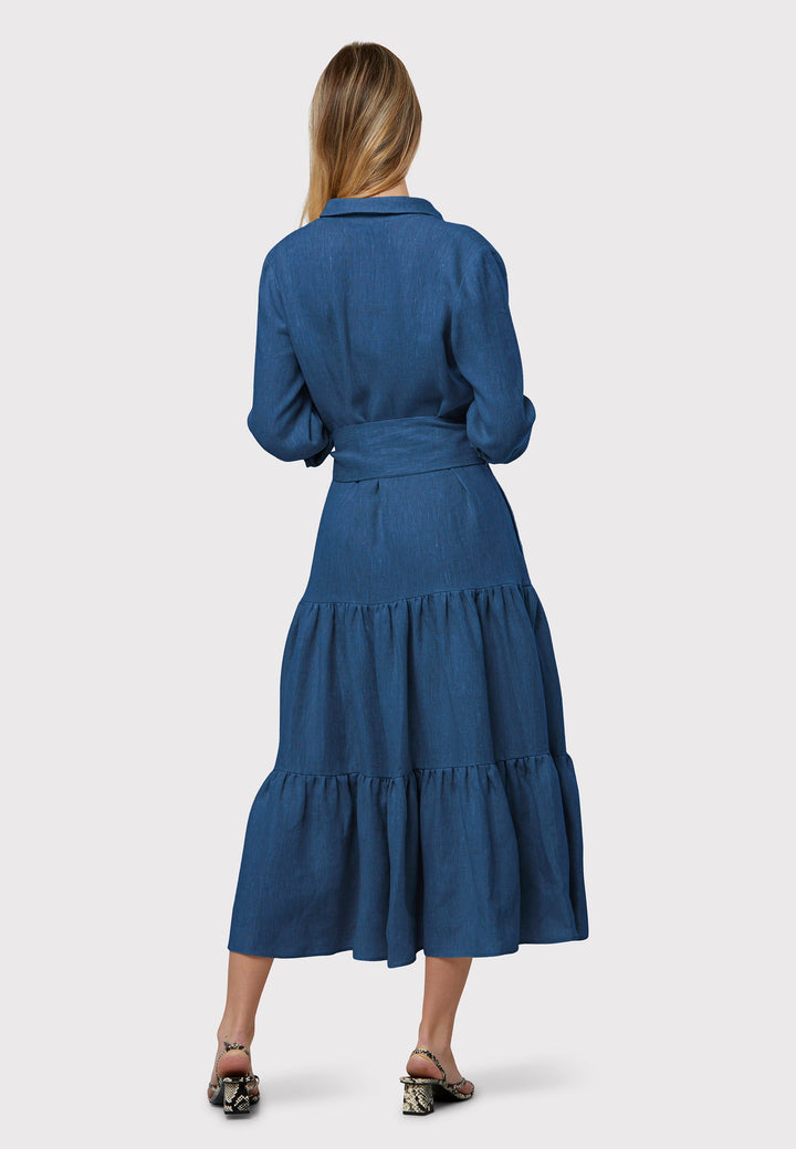 Adele Borage Blue Dress, crafted in a rich borage blue linen. The design showcases a classic shirt collar and a buttoned finish at the waist, ensuring a flattering silhouette that accentuates the hips. Transitioning seamlessly into a gathered tiered skirt, it exudes feminine charm. Complete with a detachable belt, practical pockets, and standard full-length shirt sleeves, this dress is versatile and perfect for any occasion.