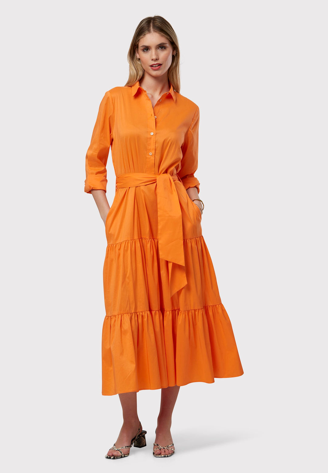 Adele Orange Dress, crafted in poplin in a vibrant orange hue. The design features a classic shirt collar and a button finish to the waist, ensuring a flattering fit that accentuates the hips. The dress gracefully transitions into a gathered tiered skirt, adding a touch of femininity. Complete with a detachable belt, practical pockets, and standard full-length shirt sleeves, this dress will take you anywhere.