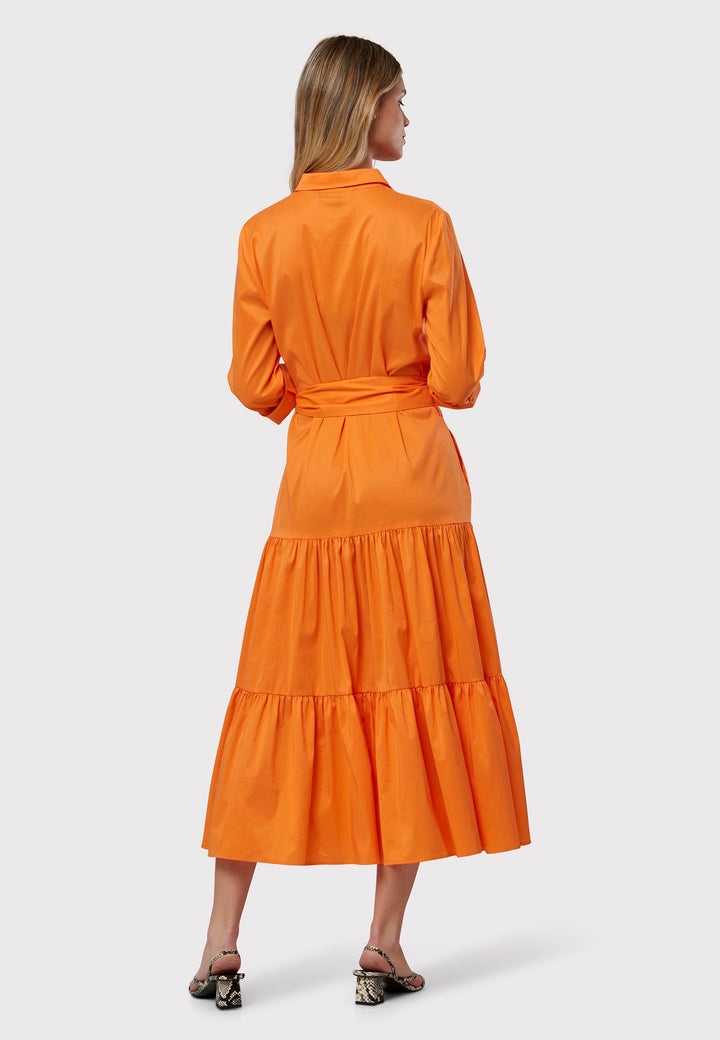 Adele Orange Dress, crafted in poplin in a vibrant orange hue. The design features a classic shirt collar and a button finish to the waist, ensuring a flattering fit that accentuates the hips. The dress gracefully transitions into a gathered tiered skirt, adding a touch of femininity. Complete with a detachable belt, practical pockets, and standard full-length shirt sleeves, this dress will take you anywhere.