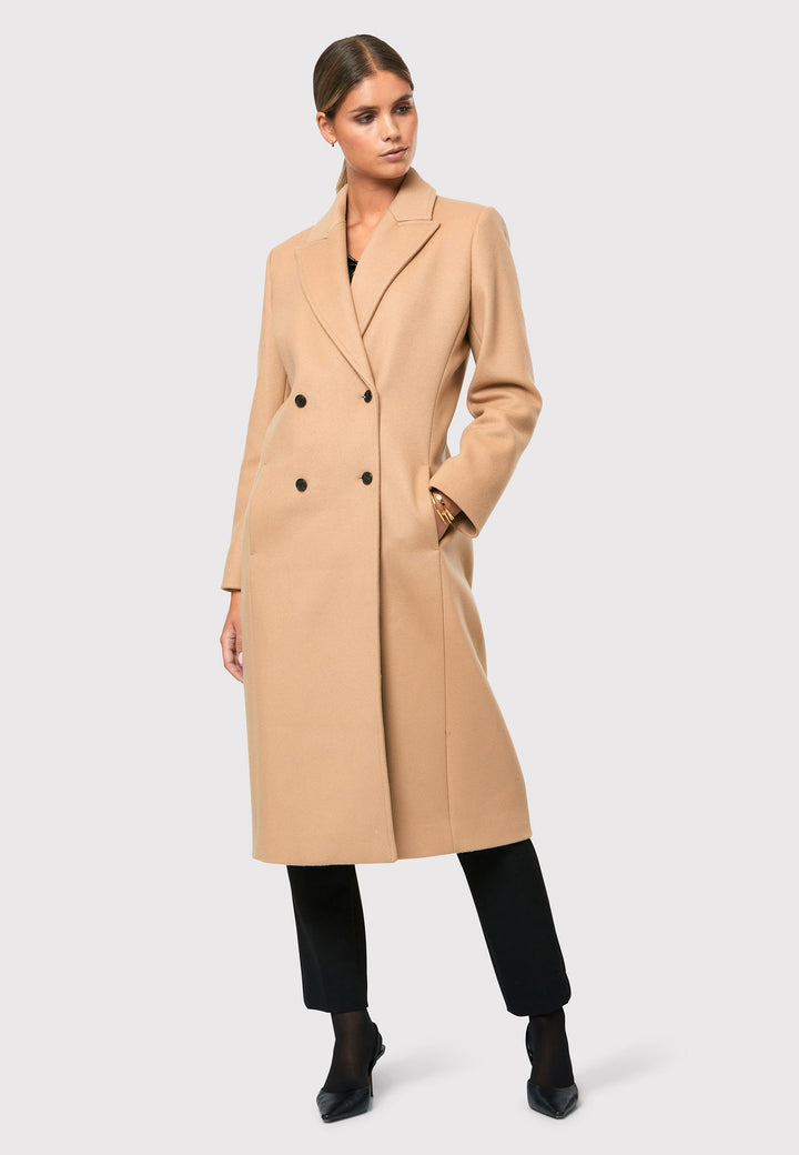 classic double-breasted coat silhouette adorned with four elegant brown horn buttons, perfectly complementing the sand color. The coat features a sleek collar and revere design. Crafted from luxurious melton fabric, it falls gracefully below the knee, enveloping you in both comfort and sophistication. 