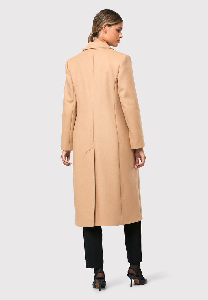 classic double-breasted coat silhouette adorned with four elegant brown horn buttons, perfectly complementing the sand color. The coat features a sleek collar and revere design. Crafted from luxurious melton fabric, it falls gracefully below the knee, enveloping you in both comfort and sophistication. 