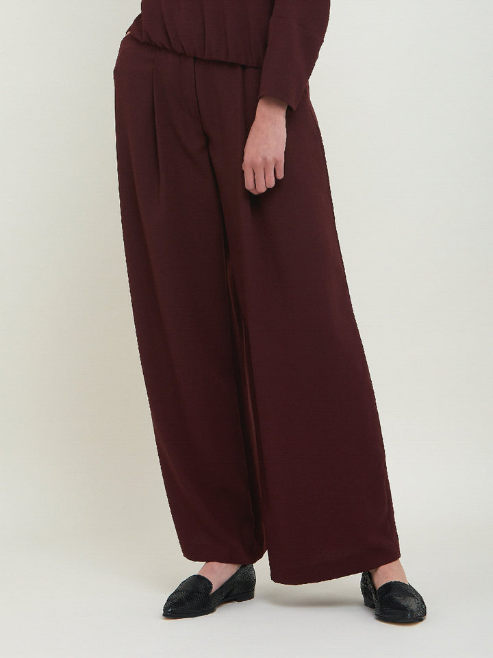 Charlize, a wide-leg palazzo pant. Crafted in a satin back crepe in rich burgundy. Features deep pleats, sharp front crease & side pockets. A high-waisted trouser made to take you from desk to dinner with a relaxed & refined silhouette. Style with flats or dress up with heels.