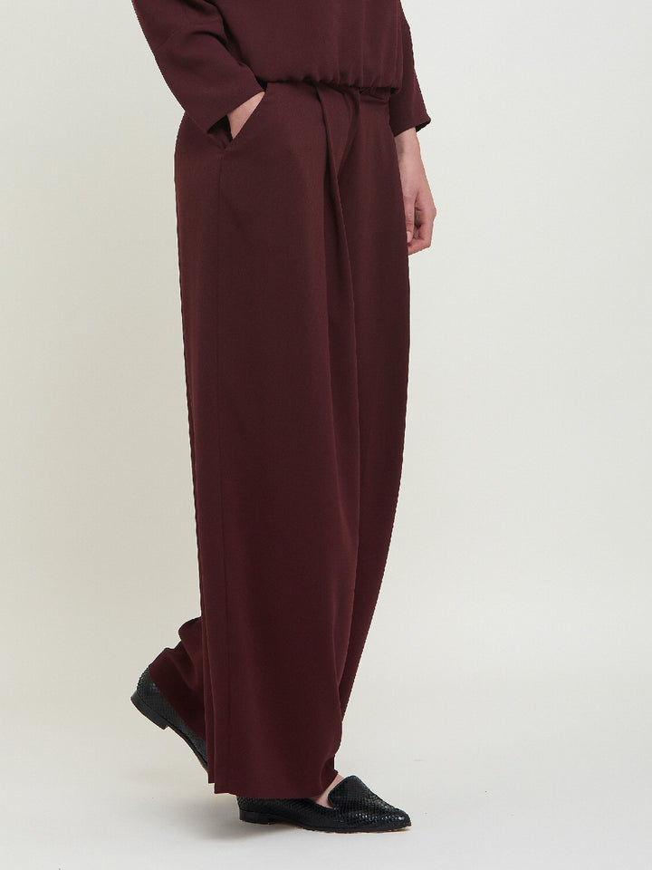 Charlize, a wide-leg palazzo pant. Crafted in a satin back crepe in rich burgundy. Features deep pleats, sharp front crease & side pockets. A high-waisted trouser made to take you from desk to dinner with a relaxed & refined silhouette. Style with flats or dress up with heels.