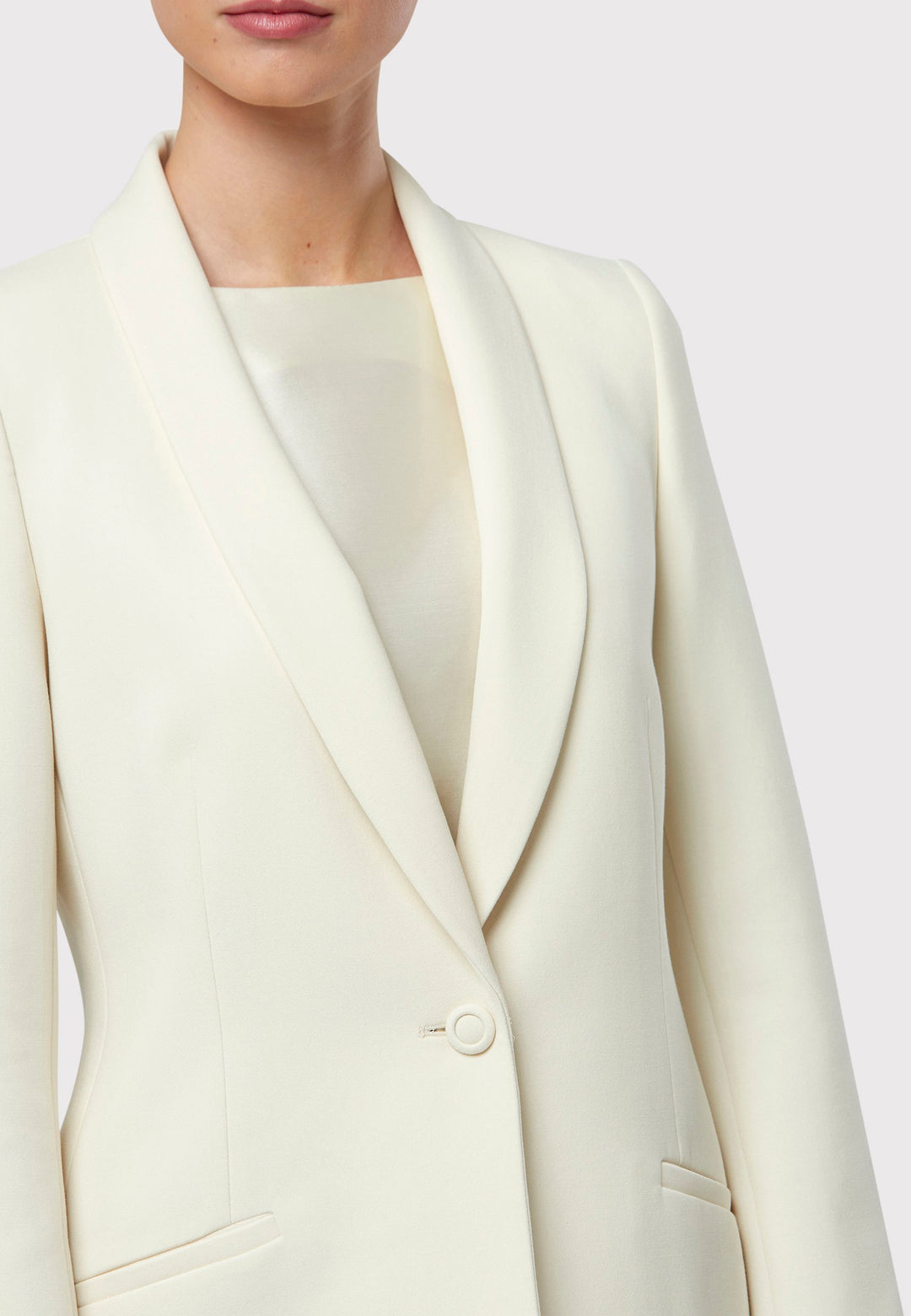 Darcie Ivory Jacket, a cornerstone for the discerning minimalist. This impeccably tailored tuxedo jacket exudes timeless elegance, featuring a seamless shawl collar and welt pockets. The semi-fitted silhouette is enhanced by a single button closure, ensuring a flattering and sophisticated look. Expertly crafted from our signature tricotine, it promises comfort and ease with just the right amount of stretch. For a polished tuxedo look, pair it with the matching Jill Ivory Trouser.