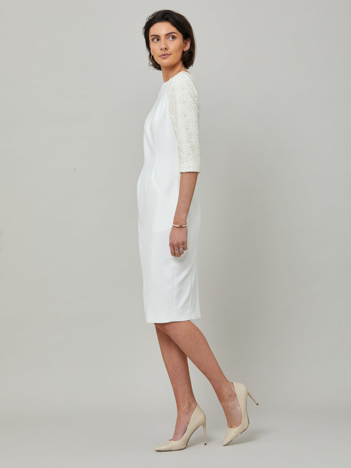 side picture of model wearing Juliet, A form-flattering, body-skimming silhouette in iconic white. Features a 3/4 sleeve with lace overlay. The skirt falls to a demure finish below the knee. Attending a summer wedding? Mother of the bride? Heading to the races? This is the dress for you.