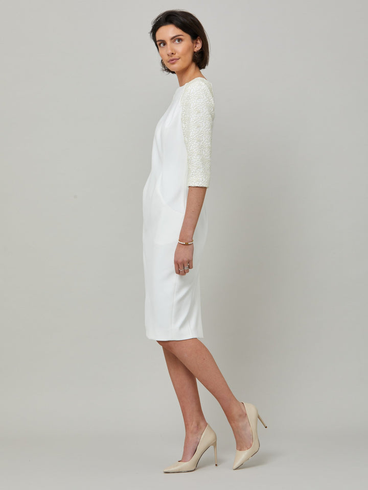 side picture of model wearing Juliet, A form-flattering, body-skimming silhouette in iconic white. Features a 3/4 sleeve with lace overlay. The skirt falls to a demure finish below the knee. Attending a summer wedding? Mother of the bride? Heading to the races? This is the dress for you.