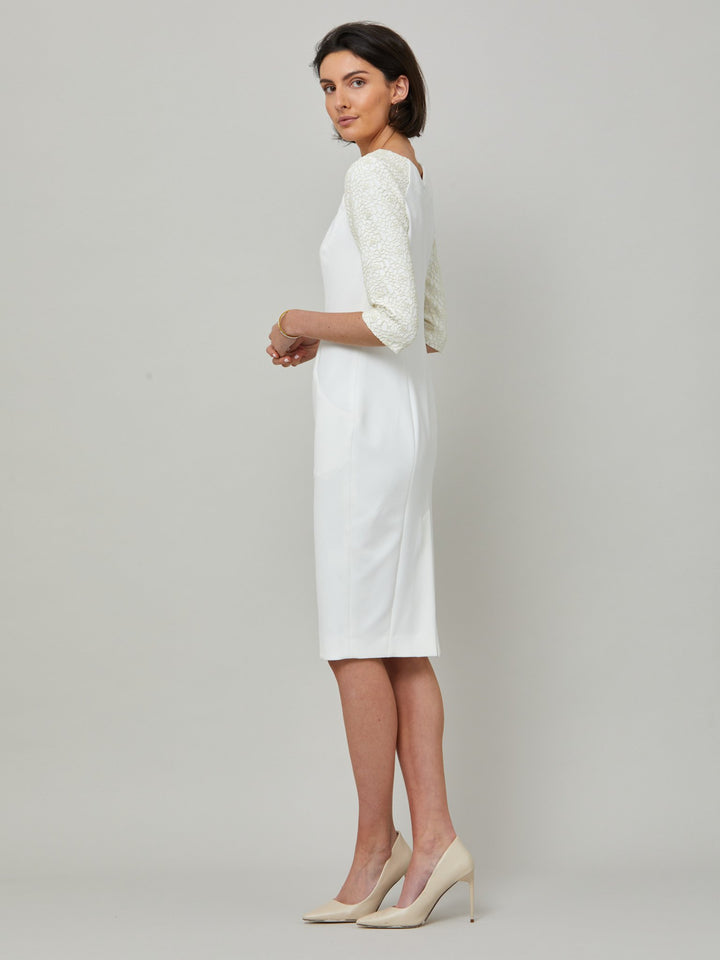 side picture of model wearing Juliet, A form-flattering, body-skimming silhouette in iconic white. Features a 3/4 sleeve with lace overlay. The skirt falls to a demure finish below the knee. Attending a summer wedding? Mother of the bride? Heading to the races? This is the dress for you.