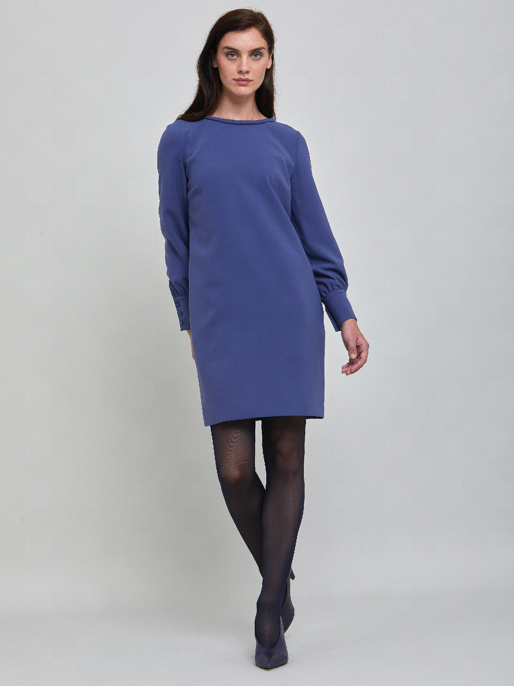 Rachel, a contemporary form-flattering shift dress. Crafted in a juniper blue tricotine with a hint of stretch. The round neck features an elegant plait trim & the easy-fitting sleeve softly gathers into a buttoned cuff. The skirt skims the knee. Work, Wine, or Dine in the Rachel Dress.
