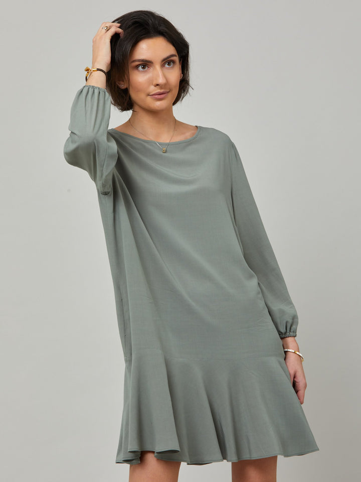 Model wearing Sheila, a great everyday dress. Crafted in our fluid lichen green viscose crepe. A flirty knee-grazing shift dress with a soft elasticated cuff sleeve that falls to a deep frill hem with a jewel neckline and key-hole back detail. For everyday comfort style with trainers. Elevate your mood with this fun piece.
