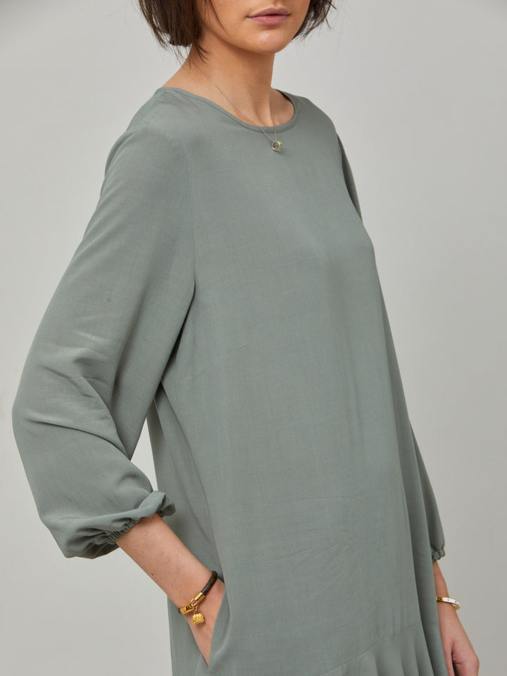 detail of Sheila, a great everyday dress. Crafted in our fluid lichen green viscose crepe. A flirty knee-grazing shift dress with a soft elasticated cuff sleeve that falls to a deep frill hem with a jewel neckline and key-hole back detail. For everyday comfort style with trainers. Elevate your mood with this fun piece.