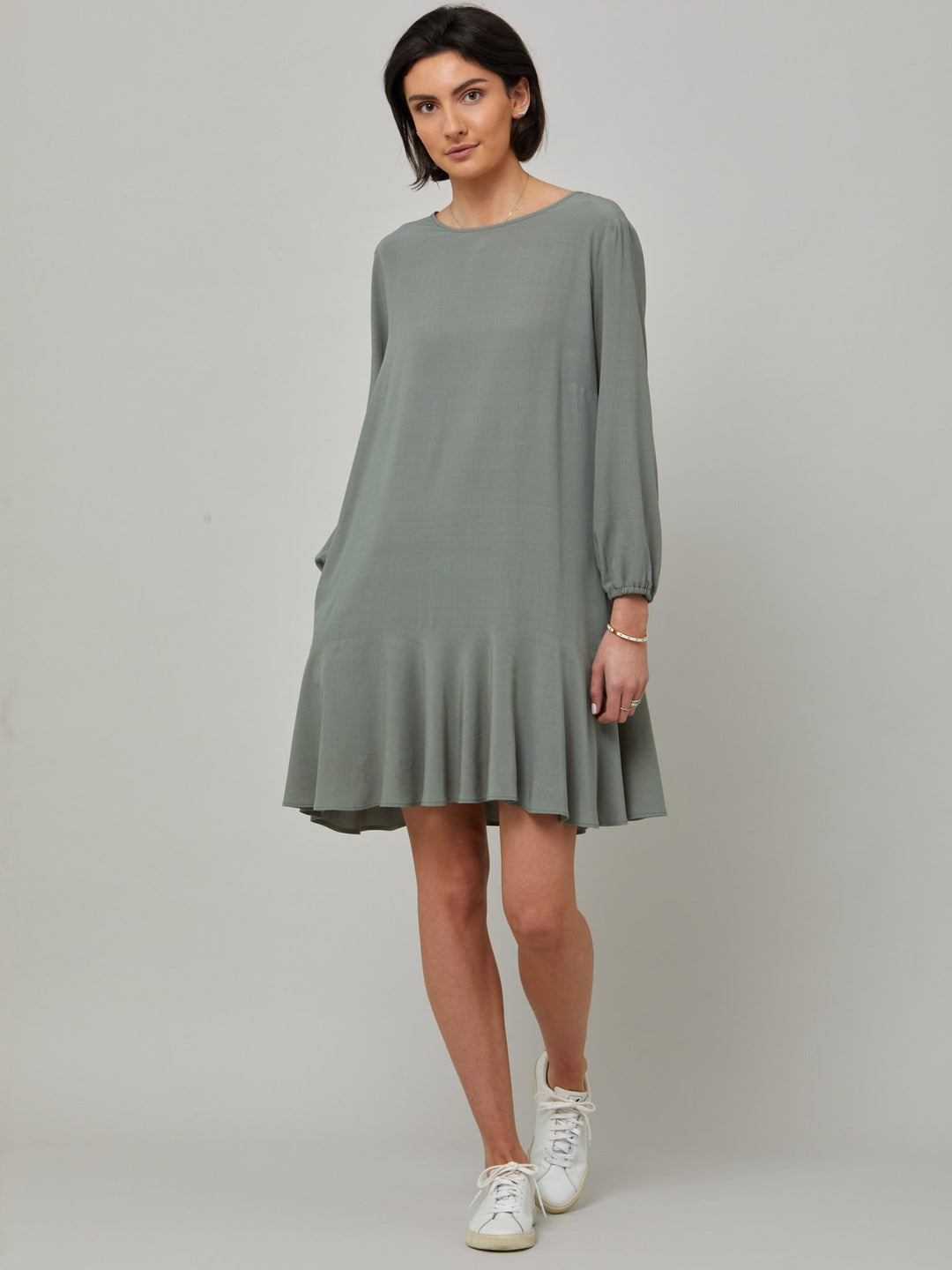front picture of model wearing Sheila, a great everyday dress. Crafted in our fluid lichen green viscose crepe. A flirty knee-grazing shift dress with a soft elasticated cuff sleeve that falls to a deep frill hem with a jewel neckline and key-hole back detail. For everyday comfort style with trainers. Elevate your mood with this fun piece.