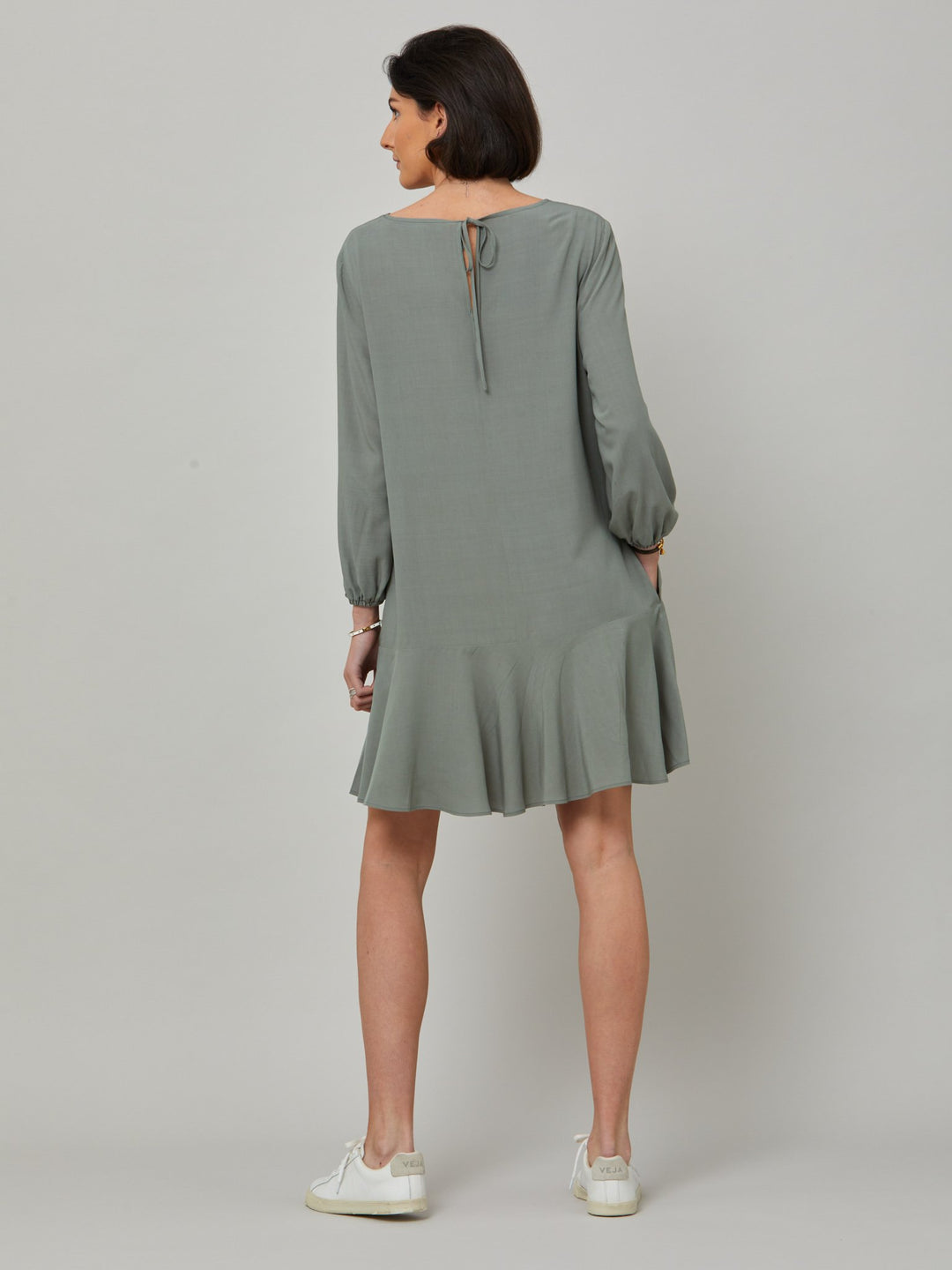 back picture of model wearing Sheila, a great everyday dress. Crafted in our fluid lichen green viscose crepe. A flirty knee-grazing shift dress with a soft elasticated cuff sleeve that falls to a deep frill hem with a jewel neckline and key-hole back detail. For everyday comfort style with trainers. Elevate your mood with this fun piece.