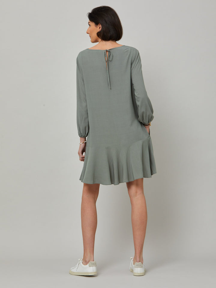 back picture of model wearing Sheila, a great everyday dress. Crafted in our fluid lichen green viscose crepe. A flirty knee-grazing shift dress with a soft elasticated cuff sleeve that falls to a deep frill hem with a jewel neckline and key-hole back detail. For everyday comfort style with trainers. Elevate your mood with this fun piece.