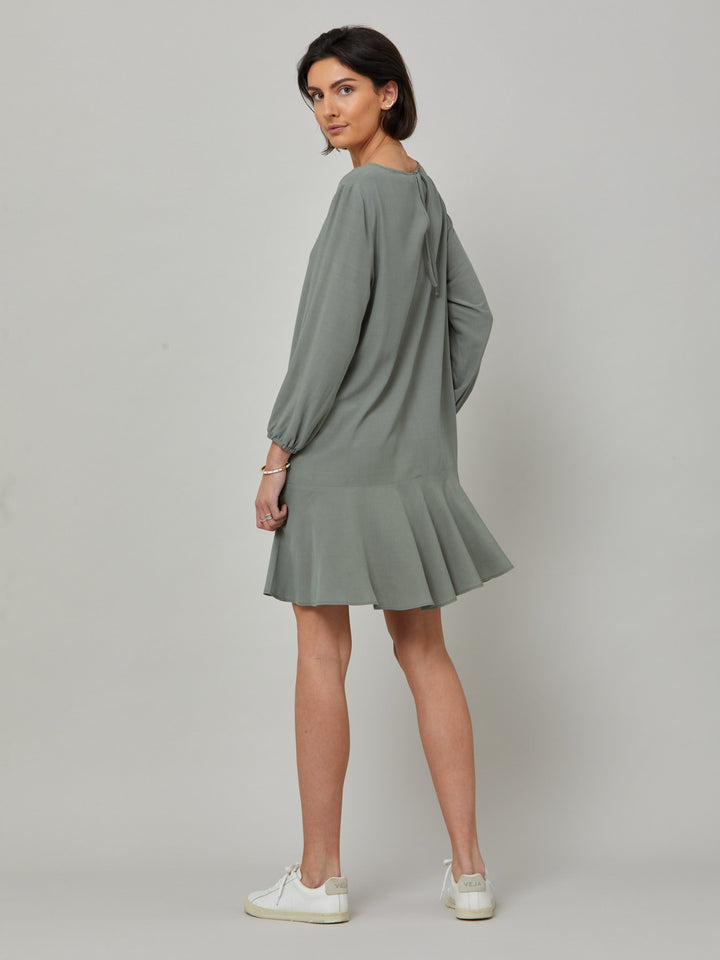 back picture of model wearing Sheila, a great everyday dress. Crafted in our fluid lichen green viscose crepe. A flirty knee-grazing shift dress with a soft elasticated cuff sleeve that falls to a deep frill hem with a jewel neckline and key-hole back detail. For everyday comfort style with trainers. Elevate your mood with this fun piece.