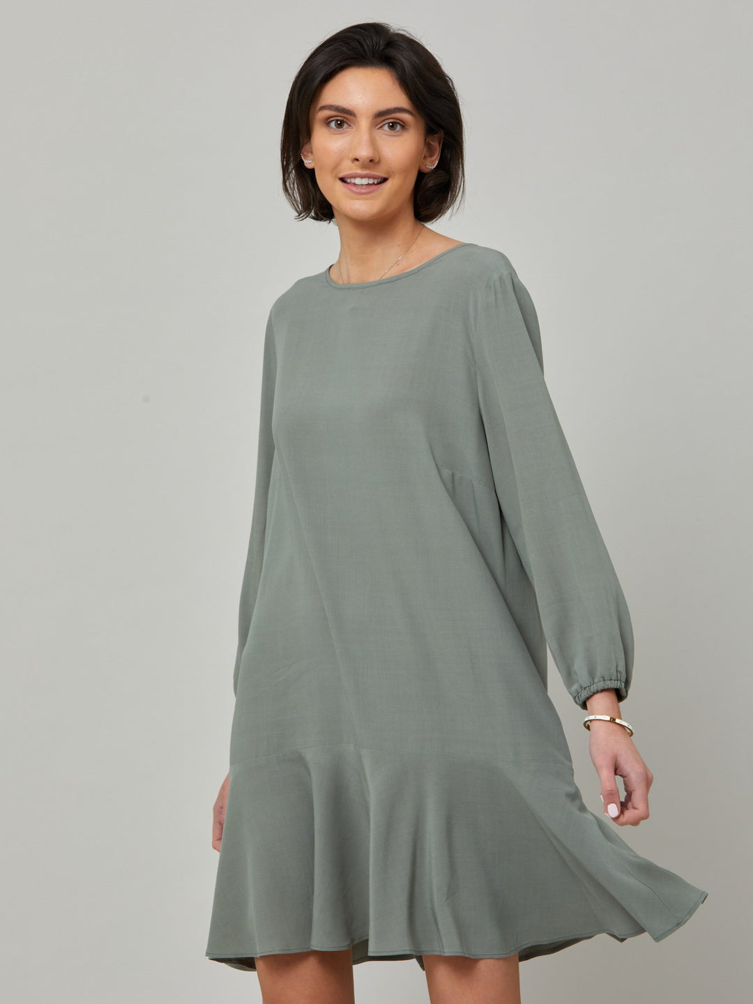 model wearing Sheila, a great everyday dress. Crafted in our fluid lichen green viscose crepe. A flirty knee-grazing shift dress with a soft elasticated cuff sleeve that falls to a deep frill hem with a jewel neckline and key-hole back detail. For everyday comfort style with trainers. Elevate your mood with this fun piece.